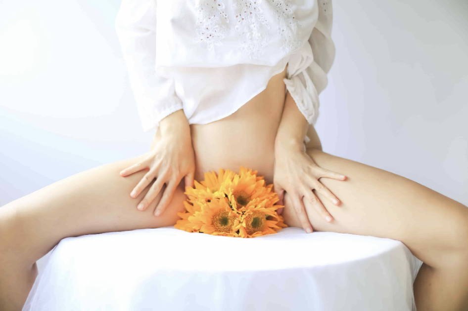 A woman sitting on a table with her legs spread, flowers covering her groin.